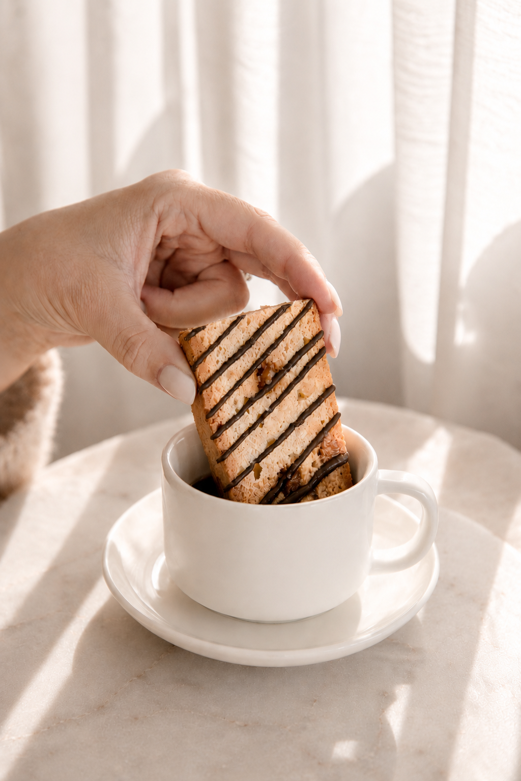A chocolate-drizzled Giorgio biscotti held over a white cup in morning light